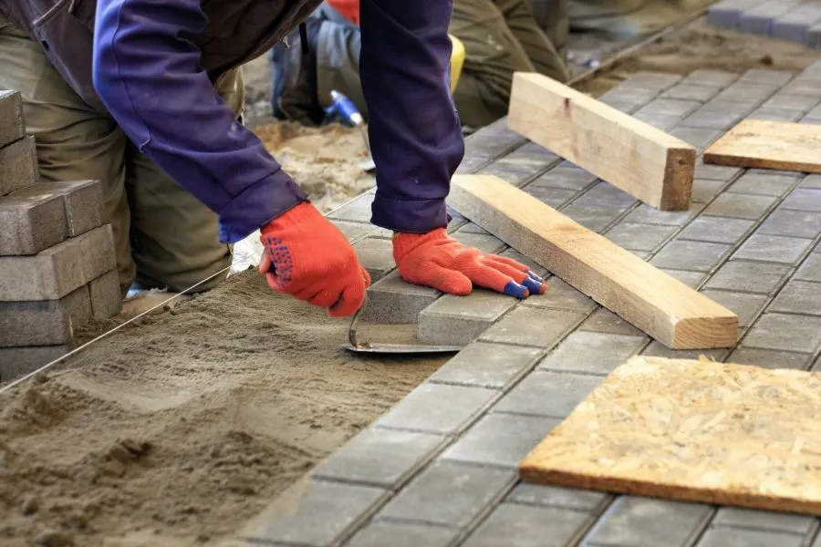 Construction worker laying paving stones on sand base with gloves and wooden spacers