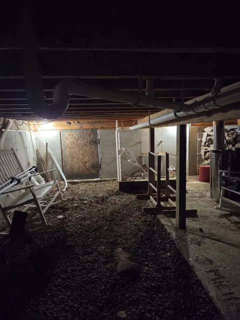 Underground construction site with exposed pipes, white chairs, and construction materials, illuminated by a single light, with a gravel floor.