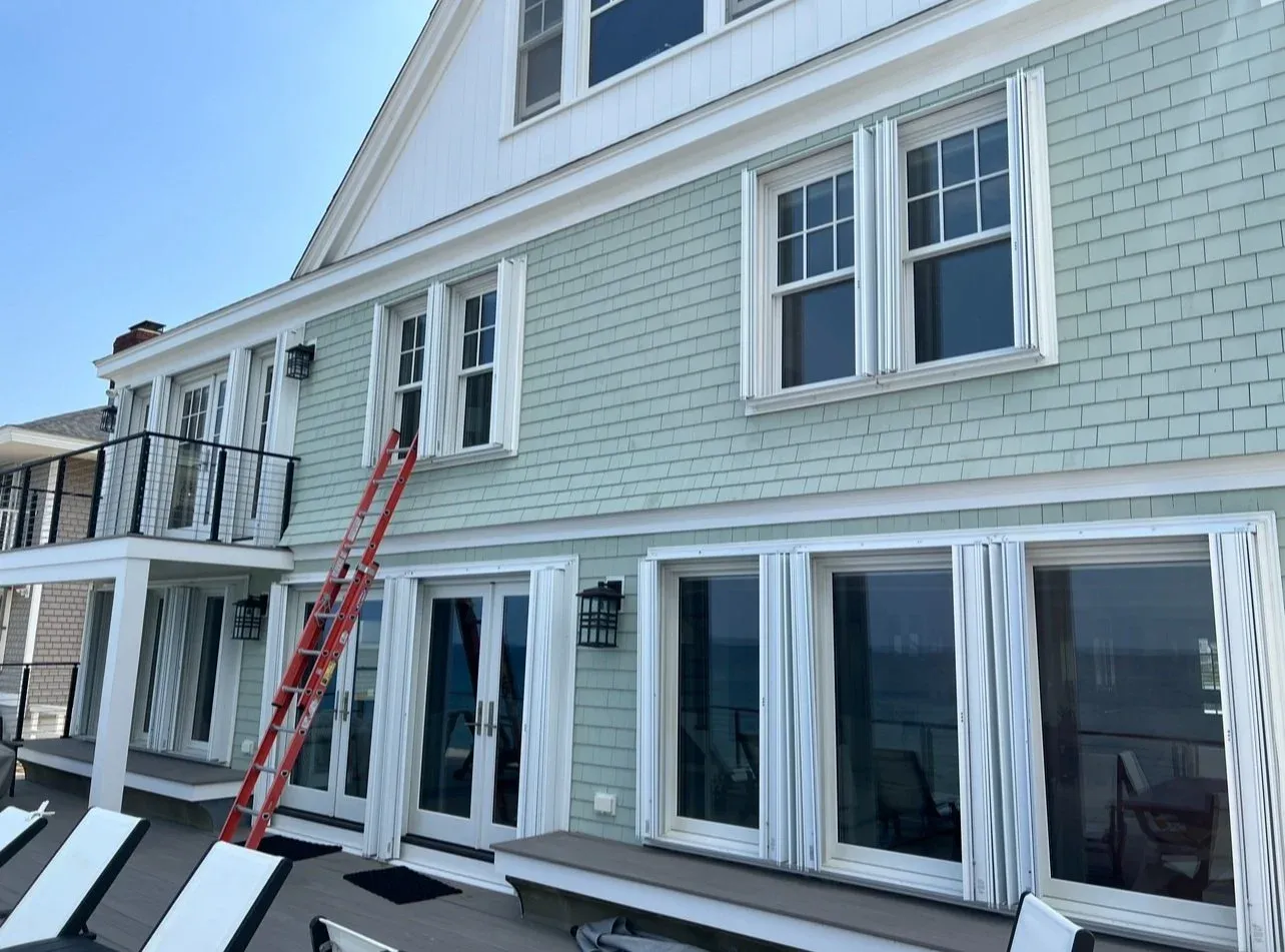 Rear view of a multi-story house with light green exterior, white trim, and multiple windows with white shutters, featuring a balcony with black railing and a red ladder leaning against the house.