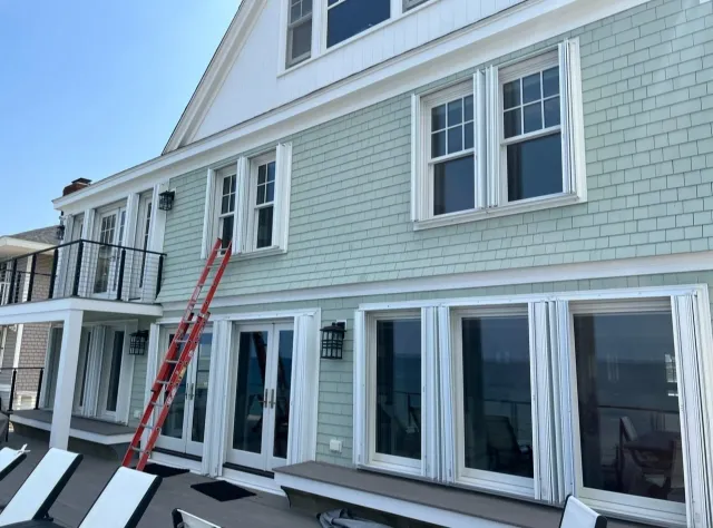 Rear view of a multi-story house with light green exterior, white trim, and multiple windows with white shutters, featuring a balcony with black railing and a red ladder leaning against the house.