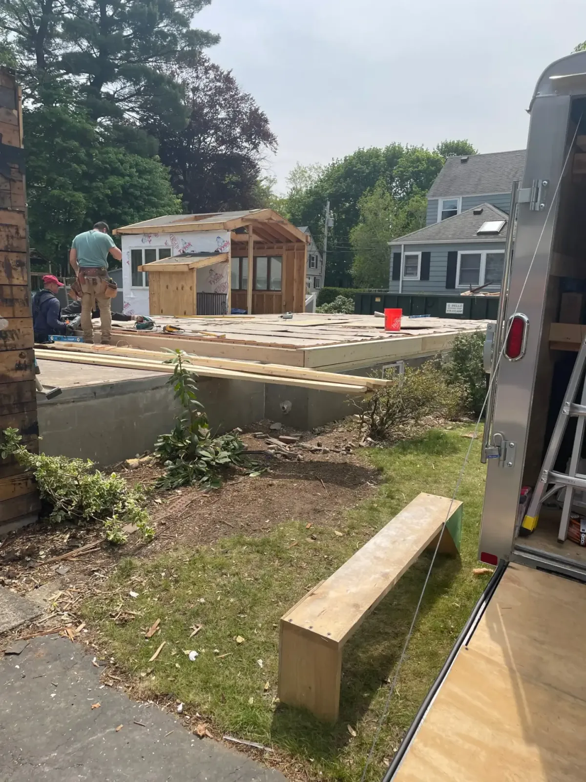 Construction site with workers framing a custom home addition in Wakefield, MA, featuring a partially built structure with wooden framing and sheathing, surrounded by greenery and tools.