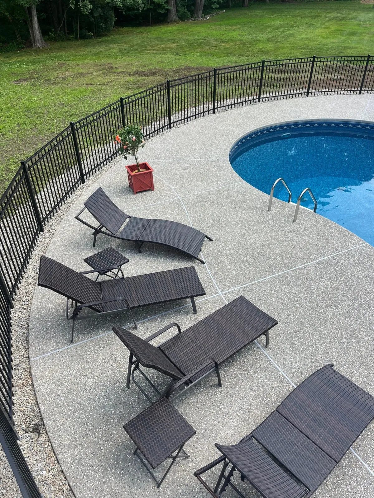 Backyard with a black metal fence, a curved in-ground swimming pool, patio furniture, potted plant, and a grassy area with trees in the background.