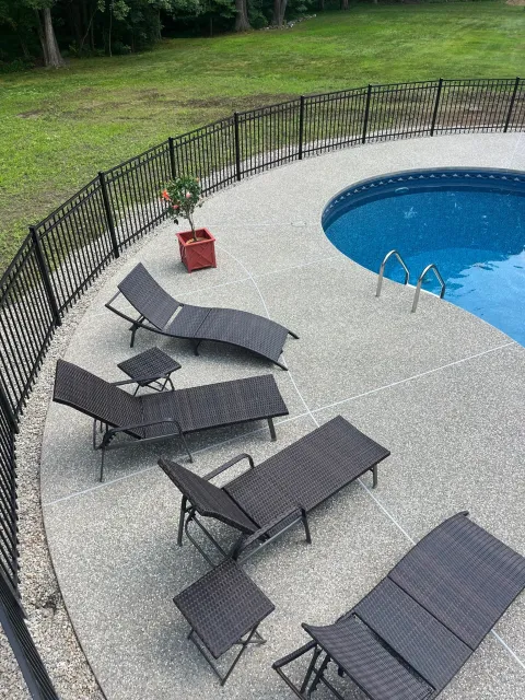 Backyard with a black metal fence, a curved in-ground swimming pool, patio furniture, potted plant, and a grassy area with trees in the background.