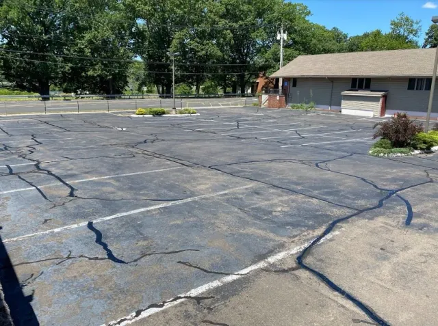 Empty parking lot with cracked asphalt and faded white parking lines, next to a grey building, with trees and a street in the background.