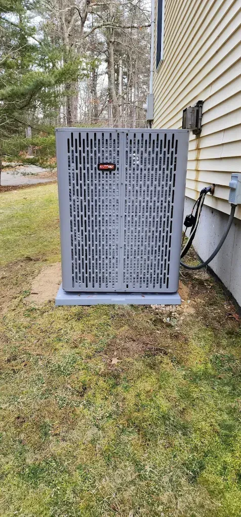 An air conditioning unit sits on a concrete pad next to a house with white siding and grass.