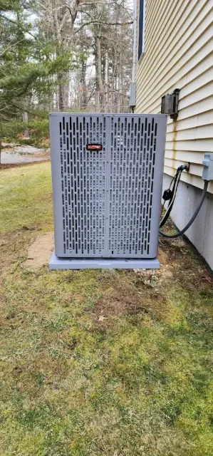 An air conditioning unit sits on a concrete pad next to a house with white siding and grass.