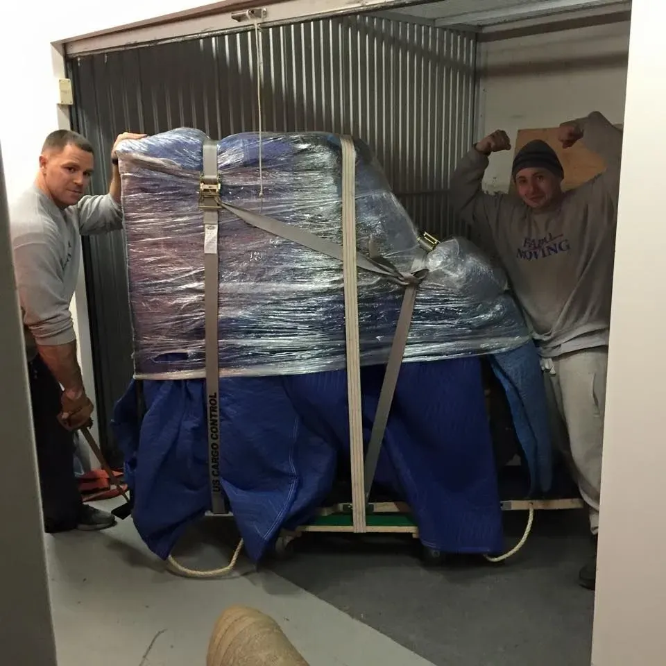 Two men moving a wrapped piano into a storage unit, one flexing his bicep.