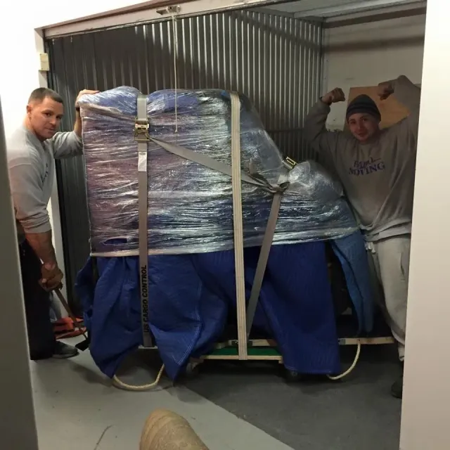 Two men moving a wrapped piano into a storage unit, one flexing his bicep.