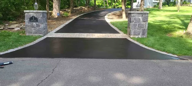 Paved driveway entrance between stone pillars, bordered by trees and green grass.