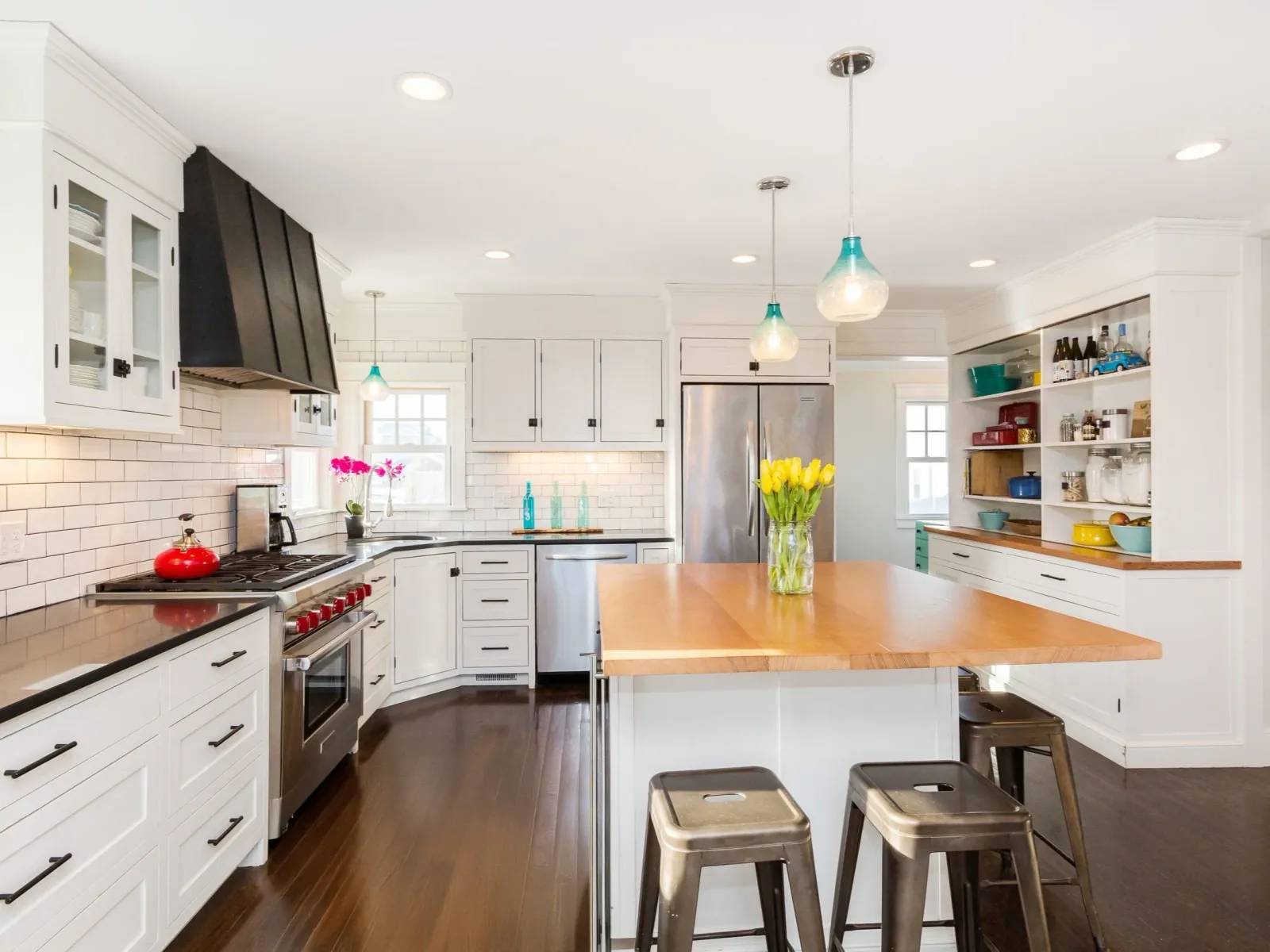 Bright, modern kitchen with white cabinets, black handles, a stainless steel refrigerator, a black range with a red kettle, and a wooden island with three metal chairs. There are decorative bottles, a potted plant, and a vase of yellow tulips on the island. Pendant lights hang from the ceiling, and there are small stools in the foreground.
