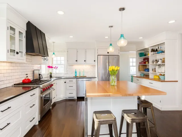 Bright, modern kitchen with white cabinets, black handles, a stainless steel refrigerator, a black range with a red kettle, and a wooden island with three metal chairs. There are decorative bottles, a potted plant, and a vase of yellow tulips on the island. Pendant lights hang from the ceiling, and there are small stools in the foreground.