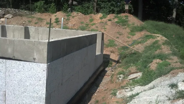 A concrete foundation wall is partially constructed in a dirt and grassy area. A pile of dirt and scattered construction materials are visible around the site.