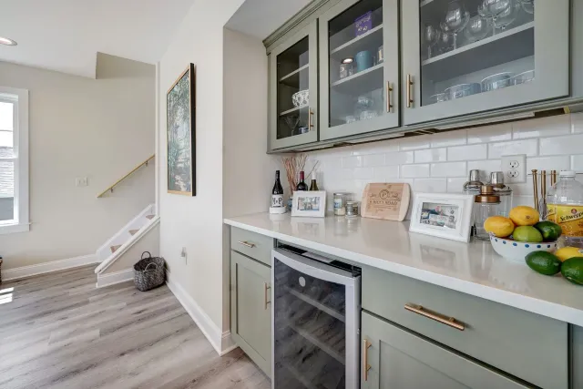 Kitchen area with green cabinets, white countertop, wine fridge, and glass-fronted cabinets.