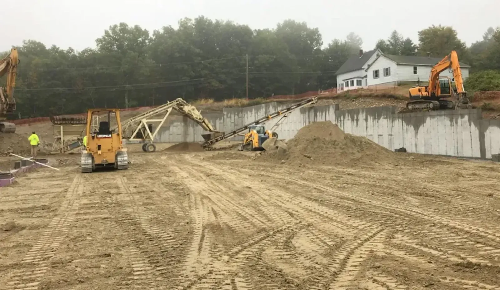 Construction site with excavators grading a dirt slope beside a concrete retaining wall and house.