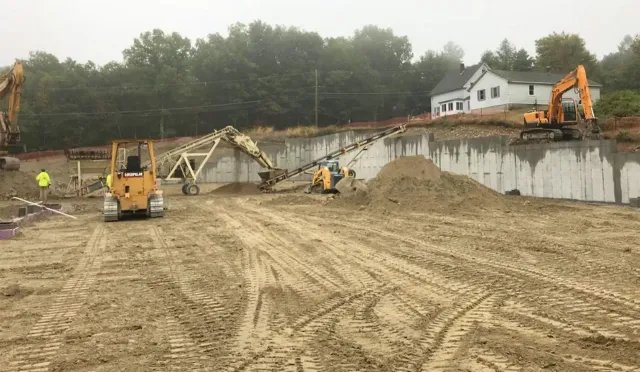 Construction site with excavators grading a dirt slope beside a concrete retaining wall and house.