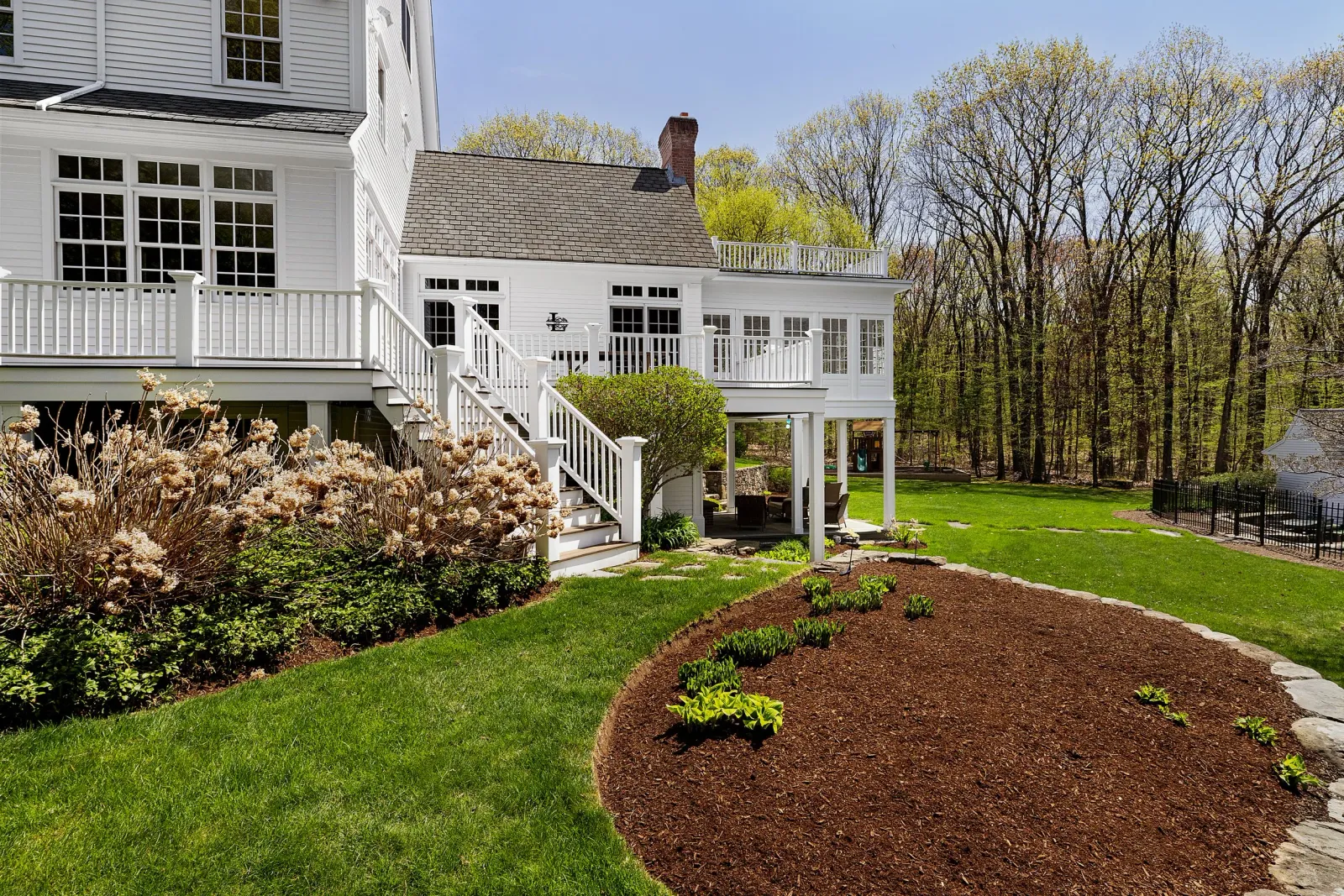 A backyard with a tiered white house with multiple decks, stairs leading to the garden, a sitting area underneath a terrace, and new plants growing in a landscaped garden with trees in the background.