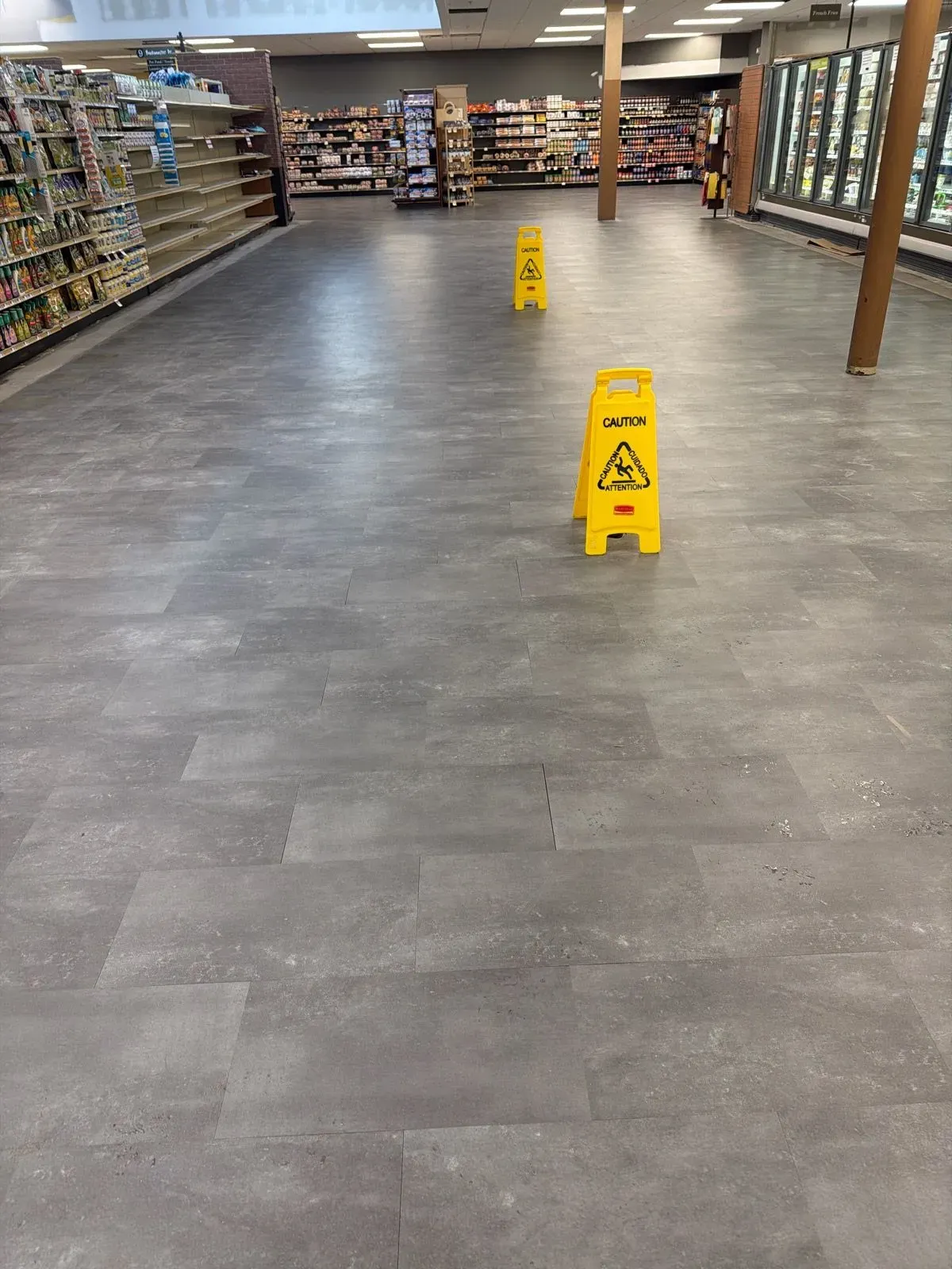 A grocery store aisle with grey stone-look vinyl flooring and two yellow caution signs positioned between empty and stocked shelves.