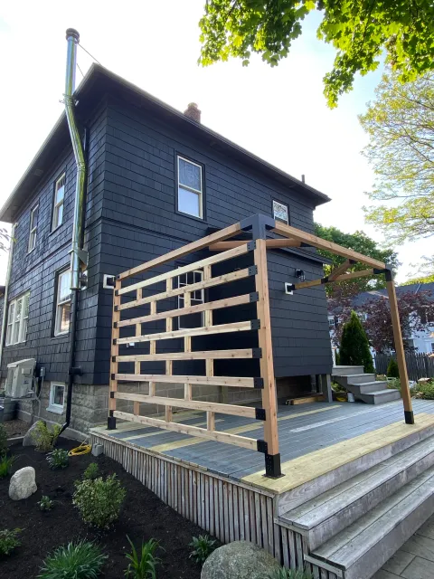 Backyard deck construction with wooden railing, stairs, and black siding house with large trees around.