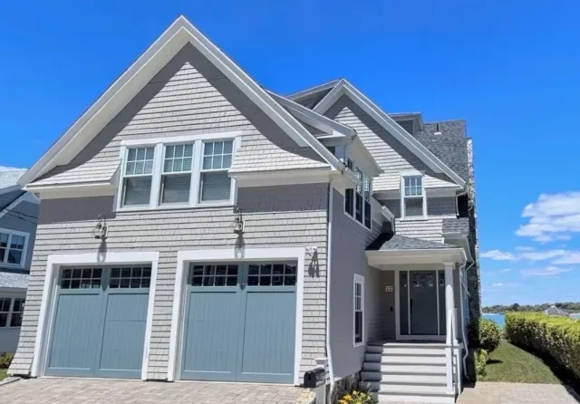 A two-story house with gray siding, a gable roof, two garage doors, and a front porch with steps, located in a neighborhood