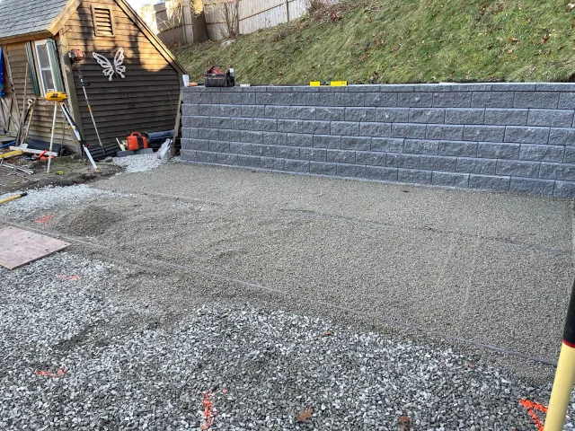 Construction site with gravel ground, a gray stone retaining wall, and a small wooden shed with butterfly decoration. Tools and construction equipment are scattered around.