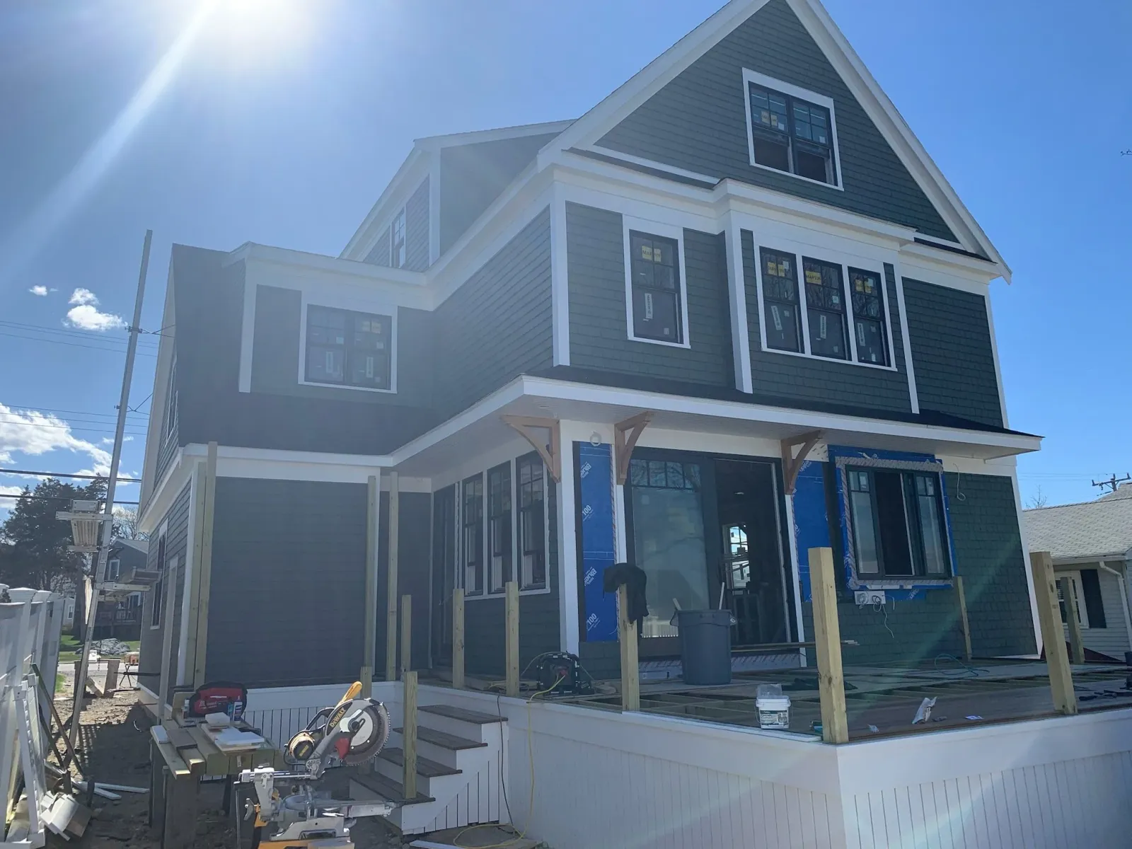 Under construction three-story house with dark green siding, multiple windows, and a front porch with steps in a sunny outdoor setting. Scituate, Norwell, Hanover, Hanson, Marshfield, South Shore. Massachusetts Local Construction Company.