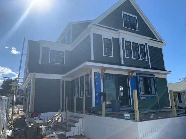 Under construction three-story house with dark green siding, multiple windows, and a front porch with steps in a sunny outdoor setting. Scituate, Norwell, Hanover, Hanson, Marshfield, South Shore. Massachusetts Local Construction Company.