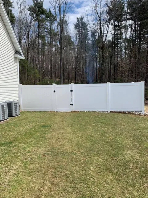 A white vinyl fence with a matching gate sits in a grassy backyard, backed by a wooded area under a partly cloudy sky.