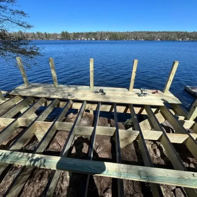 Rough wooden dock overlooking a calm blue lake under a clear sky, with a forested shoreline in the distance