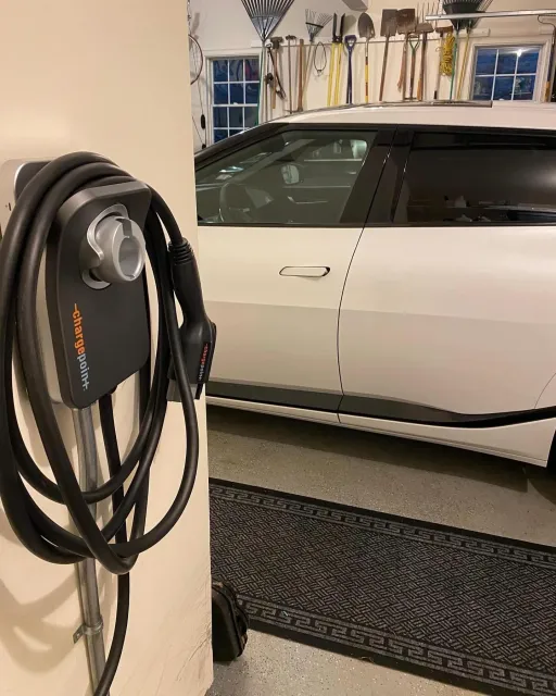 Electric vehicle charging station with a charging cable and plug in a garage, with a partial view of a white car and garage tools on the wall.