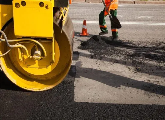 A worker spreads asphalt with a shovel while a steamroller compacts the new pavement on a road under construction.