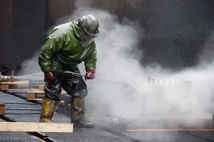 Worker in green rain gear clearing steaming asphalt with a hose on a wet construction site