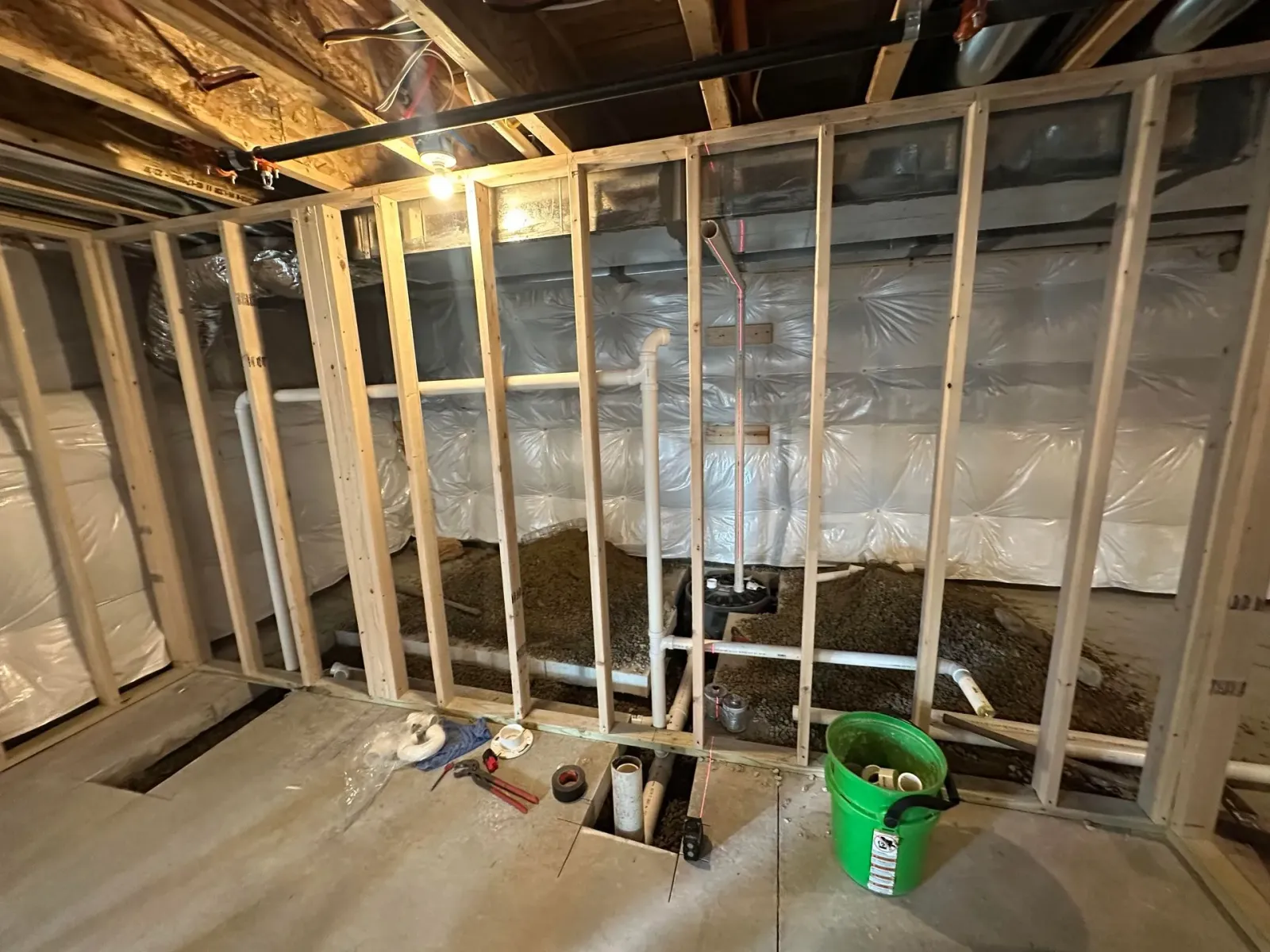 Interior view of a basement framing project. Wooden studs surround pipes and a green bucket sits on the concrete floor.