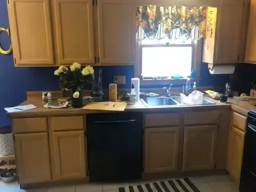 Kitchen with wooden cabinets, blue walls, sink under window, and cluttered countertop with flowers and dishes
