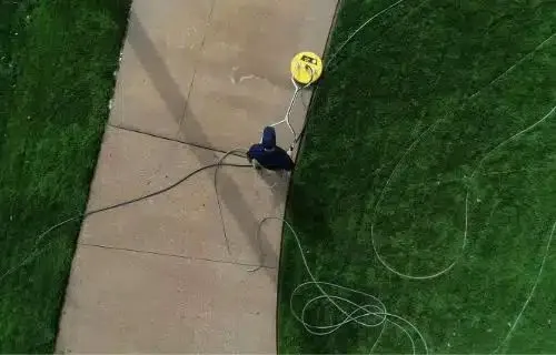 Aerial view of a person doing exterior cleaning on a concrete walkway with a yellow surface cleaner attachment. Green grass surrounds the walkway.