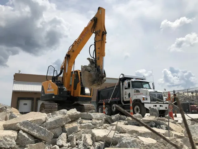 Yellow excavator loading rubble into a white dump truck, cloudy sky background.