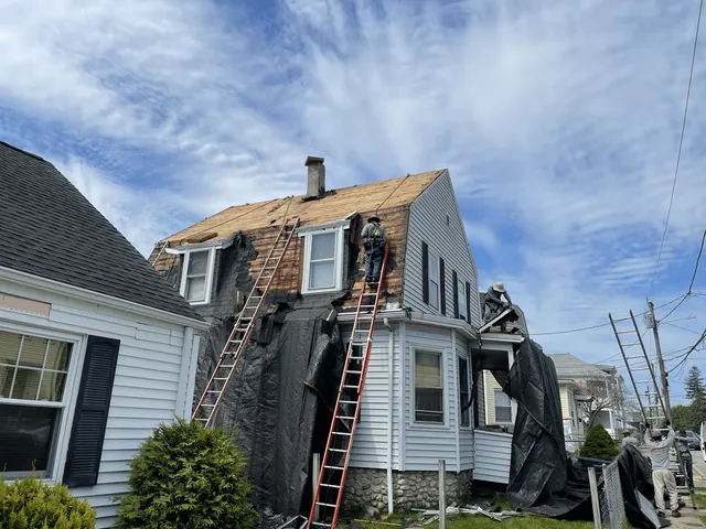 Roofing crew performing urgent tear-off and replacement work on a steep white home with safety ladders and protective tarps in place in Rhode Island.