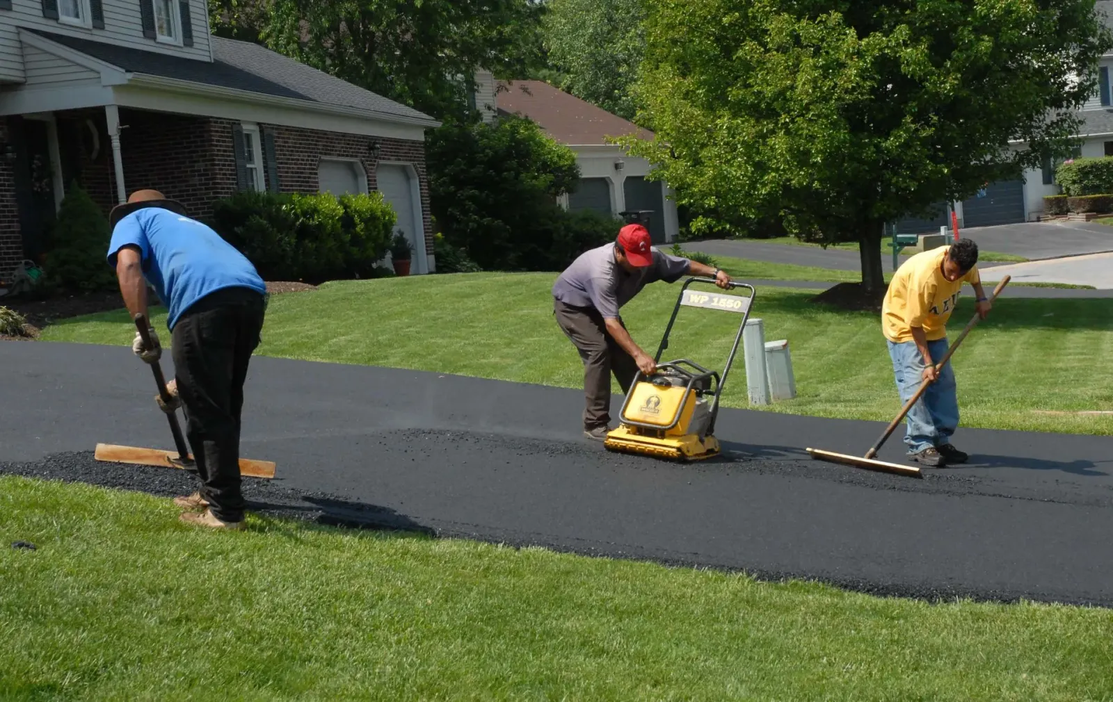 Three workers paving a driveway with asphalt: one raking, one compacting, and one using a rake.