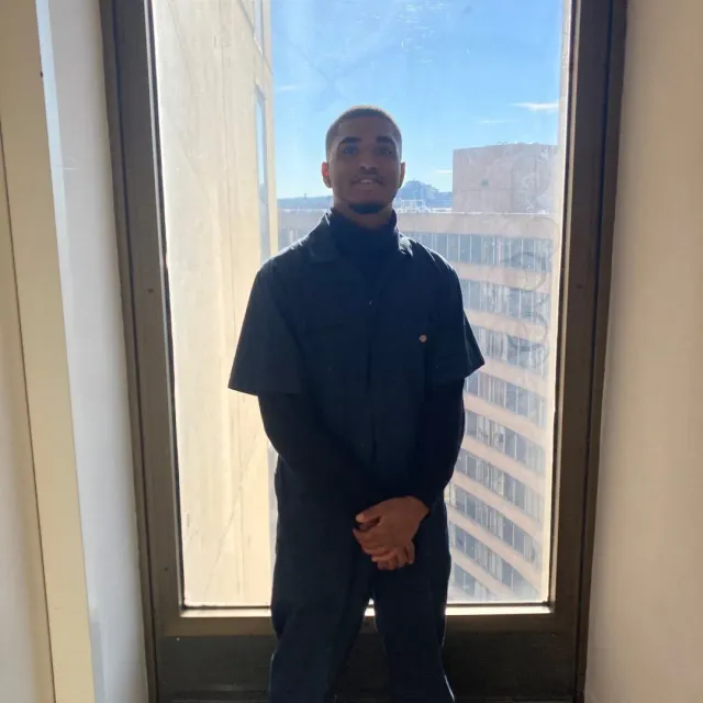 Man in black turtleneck and jumpsuit standing in front of a window, city view.
