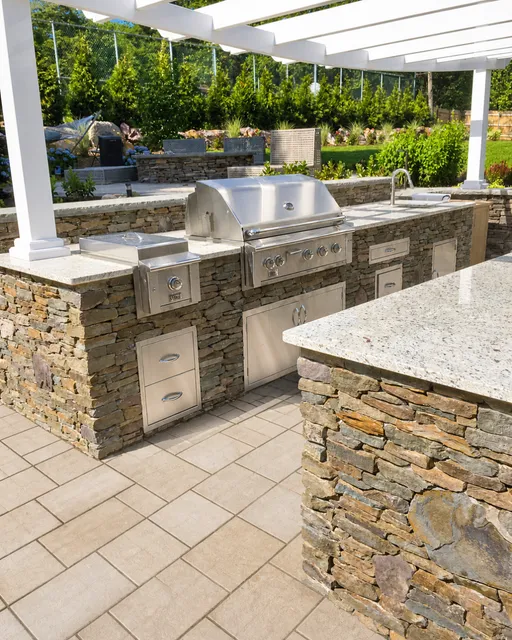Stone outdoor kitchen with stainless grill beneath a white pergola