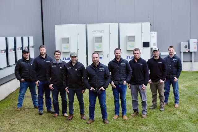 Group of people standing in front of large industrial units, wearing dark jackets on a grassy field
