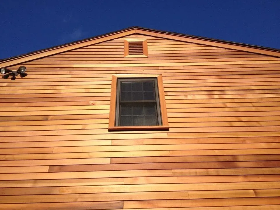 Close-up view of a house's exterior with horizontal wooden siding, a single window, and a small attic vent under a clear blue sky.