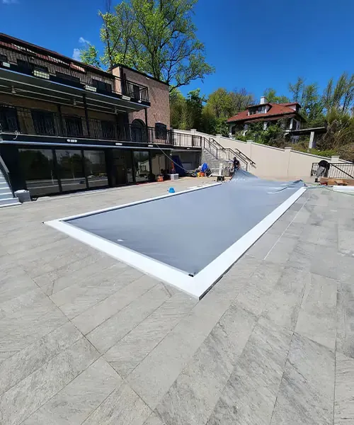 A rectangular pool with a gray cover is set in a spacious, tiled backyard patio next to a multi-story brick building, with trees and another house visible in the background under a clear blue sky.