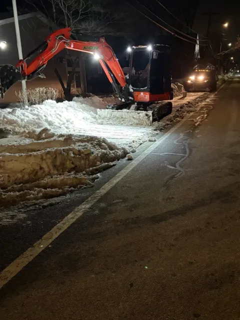 Red excavator clearing snow from a roadside at night, with a truck and headlights nearby