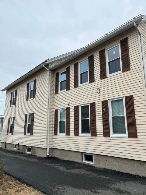 Two-story beige apartment building with brown shutters beside a paved road under a cloudy sky