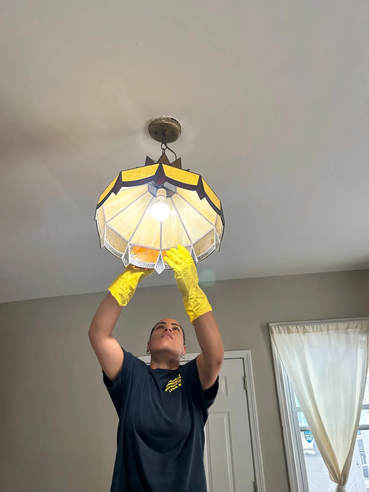 A woman in a black shirt and yellow gloves is installing or repairing a stained glass ceiling lamp in a room with white walls and a window with light-colored curtains.