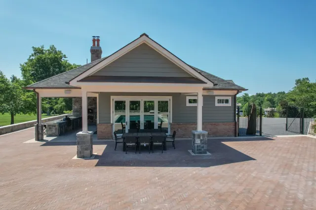 Backyard patio with brick pavers, outdoor dining table and chairs, sliding glass doors, and a built-in outdoor kitchen under a covered porch area. In the background, there are trees and a clear blue sky.