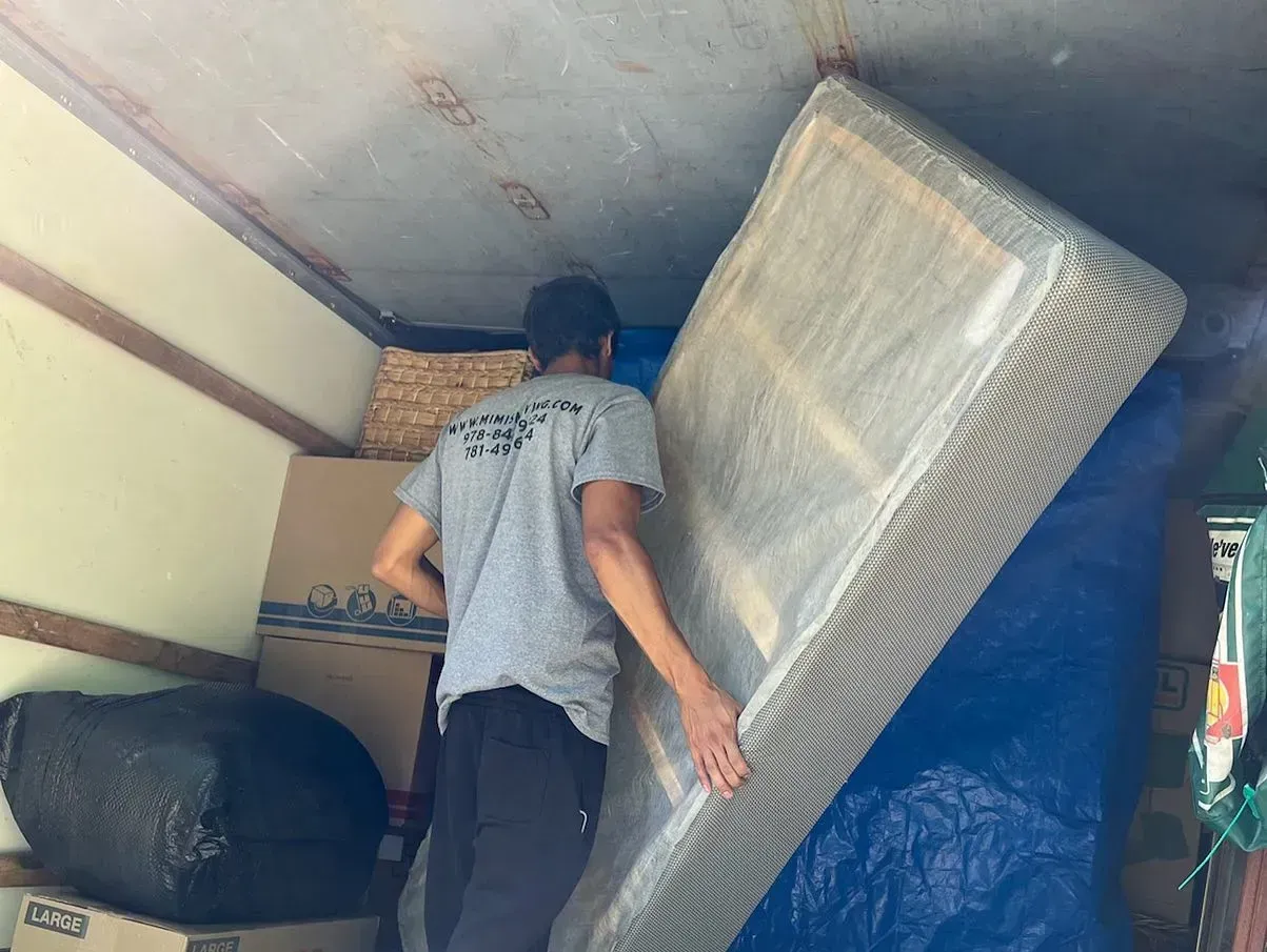 Man loads a mattress into a truck. The mattress is wrapped in plastic and leans against a blue tarp.