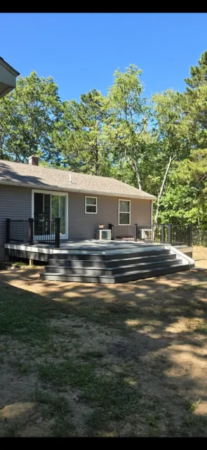 Modern residential house with gray siding and multi-level black deck surrounded by tall trees on sunny day