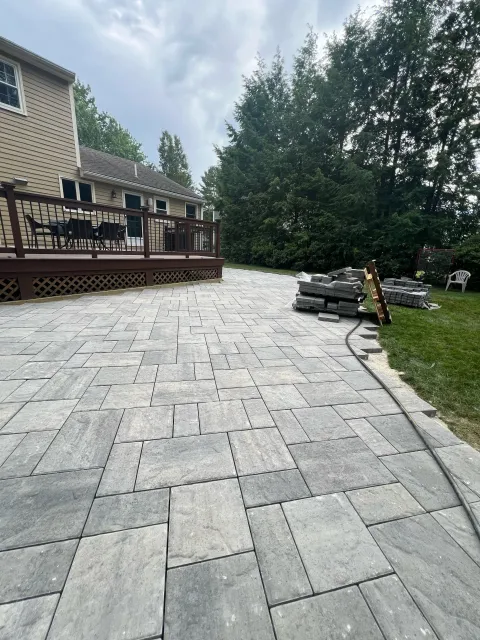 Newly installed gray stone pavers on a backyard patio with a wooden deck, construction materials, and a garden chair, surrounded by trees and residential houses.