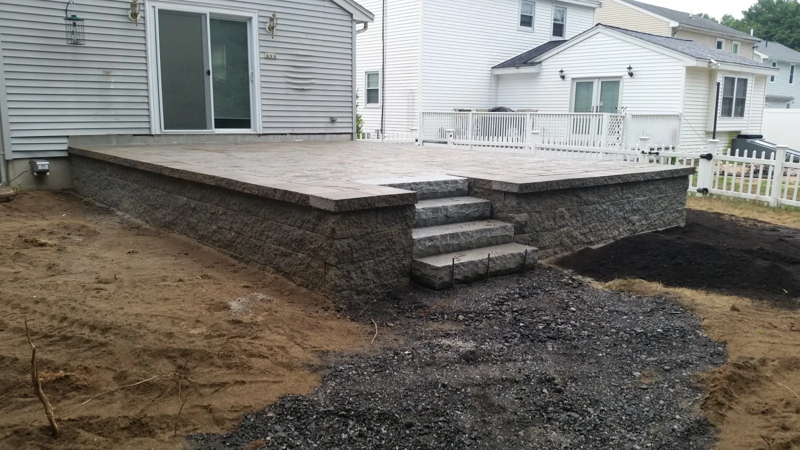 A newly constructed stone patio with steps leading down to a dirt yard, attached to a house with white siding and large sliding glass door.